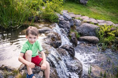 Genç bir çocuk nehir kıyısında oturuyor, yukarıdan manzaralı. Sıcak yaz ya da bahar günü. Tatlı beyaz çocuk suyla oynuyor..