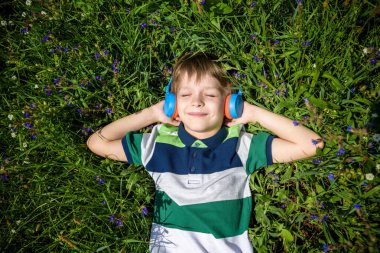 Kollarını nbaşının altında gülümseyen çocuk yeşil çimenlerin üzerinde güneşin altında yatıyordu ve okuldan sonra müzik dinliyordu. Fotoğraf yukarıdan. Doğa müziği dinlenmek kavramı.