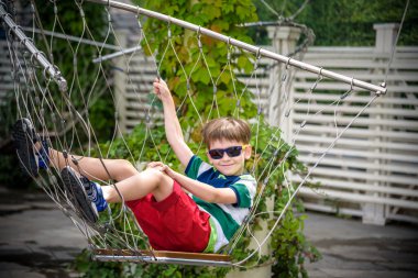 Yaz tatilinde zincirli salıncakta oturan sevimli çocuk. Çocuk rahat ve güneş gözlüğü takarak mutlu. Şehir konseptinde çocuklar için aktif yaz tatili.