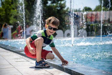 Küçük çocuk havuz kenarındaki meydanda güneşli yaz gününde çeşmede su jetleri ile oynuyor. Şehirdeki çocuklar için aktif yaz tatili.