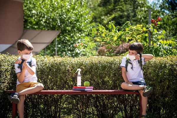 Coronavirus school reopening concept: a school boys sit on bench in front of a school wearing a face mask. Communicating trough smart watch. Covid-19, health, education, safety, and back to school.