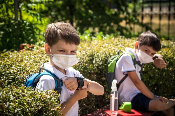 Coronavirus school reopening concept: a school boys sit on bench in front of a school wearing a face mask. Communicating trough smart watch. Covid-19, health, education, safety, and back to school.