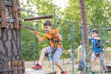 İki sevimli mutlu genç çocuk, çocuk ve kardeşi koruyucu koşum takımı içinde, karabina ve emniyet kaskları halatta, parlak güneşli bir günde yeşil yapraklı bokeh arka planda. Açık hava aktiviteleri, oyun konsepti.