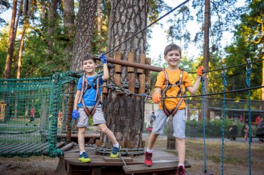 İki sevimli mutlu genç çocuk, çocuk ve kardeşi koruyucu koşum takımı içinde, karabina ve emniyet kaskları halatta, parlak güneşli bir günde yeşil yapraklı bokeh arka planda. Açık hava aktiviteleri, oyun konsepti.