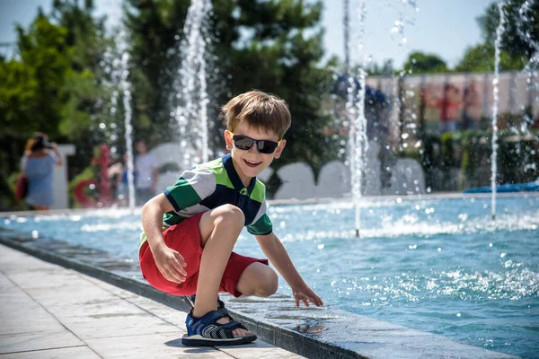 Küçük çocuk havuz kenarındaki meydanda güneşli yaz gününde çeşmede su jetleri ile oynuyor. Şehirdeki çocuklar için aktif yaz tatili.