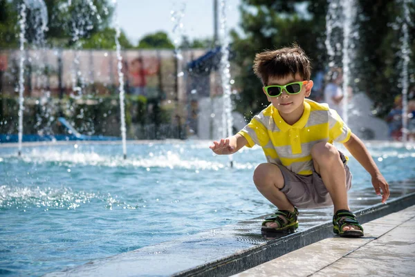 Küçük çocuk havuz kenarındaki meydanda güneşli yaz gününde çeşmede su jetleri ile oynuyor. Şehirdeki çocuklar için aktif yaz tatili.
