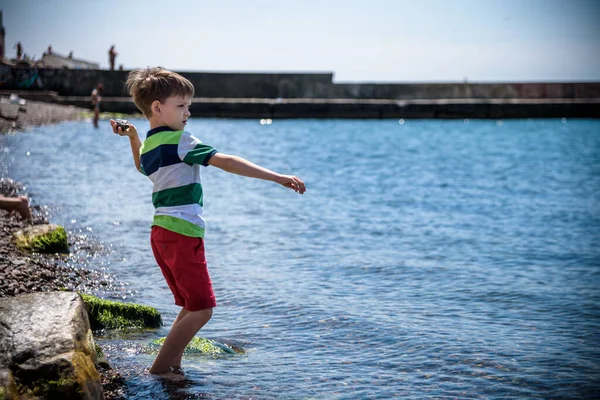 The little boy by the sea throws stones in water. Relaxation and vacation with kids. Happy childhood.