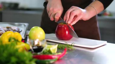 Kadın elleri, mutfakta sebzelerle çevrili büyük, sulu kırmızı bir elmayı dilimliyor. Evde taze, doğal, sağlıklı gıda hazırlama süreci.