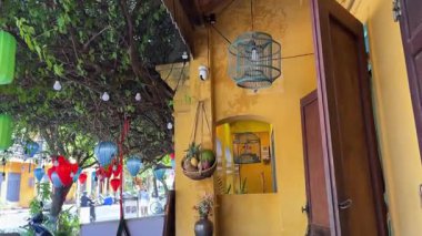 Hoi An 'daki sarı bir binadan sarkan geleneksel Vietnam ipeği fenerlerinin yakın çekimi, kısmen yeşil yapraklar ve beyaz çiçeklerle gizlenmiş. Yüksek kalite 4k görüntü