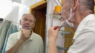 Yaşlı, İspanyol, sakallı, yakışıklı bir adam banyoda aynaya bakarak tıraş oluyor. Günlük rutin erkek hijyenik güzellik prosedürü. Adam bıyık ve sakal kesiyor.