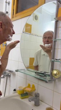 Kıdemli adam yüzünü tıraş ediyor. Yaşlı erkek, bıyığını traş makinesiyle tıraş eder. Yaşlı gri saçlı adam aynaya bakıyor, tıraş köpüğü sürüyor ve jilet kullanıyor. Deri hijyeni. Günlük vücut bakımı.