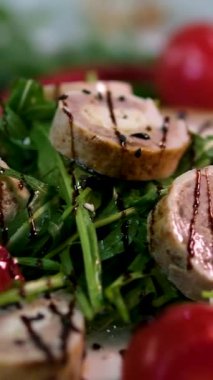 Beyaz bir tabağa yerleştirilmiş roka, sonra dilimlenmiş tavuk ve ciğer rulolarını domates dilimleriyle yerleştirilmiş olarak gösteren bir tarif. siyah ve beyaz susam ve zeytinyağı ile bitti..