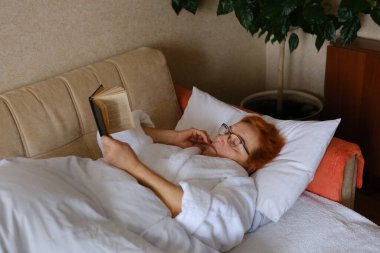 Evde yatakta kitap okuyan bir kadın. Yetişkin, cüppeli kadın yatak odasında yatakta oturuyor.