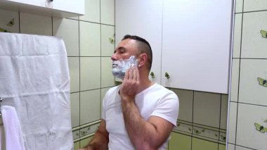 Beyaz adam, aynaya bakarak tıraş köpüğü ve fırçayla tıraş oluyor. Bir adam yüzünü duvarında kelebekler ve açık yeşil fayanslarla banyoda tıraş ediyor..