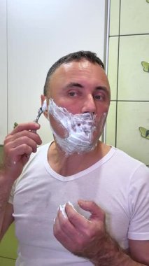 Beyaz adam, aynaya bakarak tıraş köpüğü ve fırçayla tıraş oluyor. Bir adam yüzünü duvarında kelebekler ve açık yeşil fayanslarla banyoda tıraş ediyor..