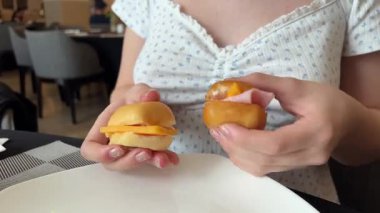 Elinde iki küçük sandviç tutan bir kız. Fast food sandviçi yiyen bir kadın. Aç genç kız fast food yiyor sağlıksız taze sandviç hamburger ve müzik dinliyor.. 