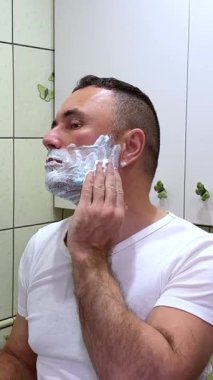 Beyaz adam, aynaya bakarak tıraş köpüğü ve fırçayla tıraş oluyor. Bir adam yüzünü duvarında kelebekler ve açık yeşil fayanslarla banyoda tıraş ediyor..