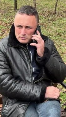 Deri ceketli bir adam sonbaharda elinde telefonla bankta oturur. Yüksek kalite 4k görüntü