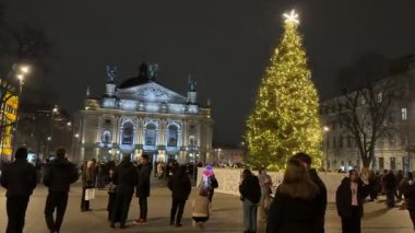 Aileler ve ziyaretçiler, Opera Binası yakınlarında neşeli bir akşamın keyfini çıkarıyorlar. İnsanlar yürürken renkli ışıklarla parlayan parlak Noel ağacı standları, sıcak bir tatil ortamı yaratıyor. Lviv Ukrayna 12.10.2025