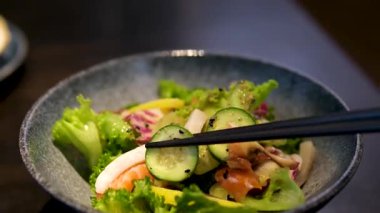 Taze sebze, tohum ve deniz ürünleriyle dolu renkli bir yemek kabından yiyecek toplayan yemek çubuklarının yakın çekimi, sağlıklı yeme ve Asya füzyon mutfağını sembolize ediyor. Yüksek kalite 4k