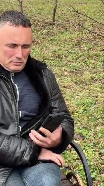 Gri saçlı orta yaşlı bir adam sonbaharda bankta oturur, telefonla konuşur, numarayı çevirir, deri ceket giyer, kot pantolon giyer, Avrupalı erkekler, siyah saçlar. Yüksek kalite 4k görüntü