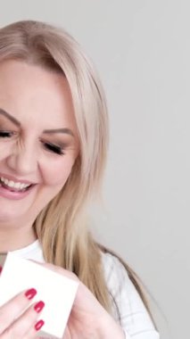 Kırk beş yaşındaki mutlu Avrupalı bir kadının elinde bir hediye kutusunu tutarken ve açarken dans ederken ve kırmızı tırnaklı beyaz kirpikleriyle anın keyfini çıkarırken gösteren canlı bir video seti..