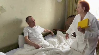 Bir kitap okuduk ve birbirimize sarıldık. Öpüşme, tablet ve yatak ağındaki yaşlı çift birlikte rahatlıyor. Aşk, emeklilikteki yaşlı adam ve kadın evdeki yatak odasında kaynaşıyorlar. Yüksek kalite 4k