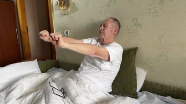 Sabah egzersizleri ellerinizi sıkar, parmaklarınızı sıkar, yumruklarınızı sallarsınız. Yetişkin bir adam yatakta hafif ısınma hareketleri yapar, kollarını yana açar. 4k görüntü