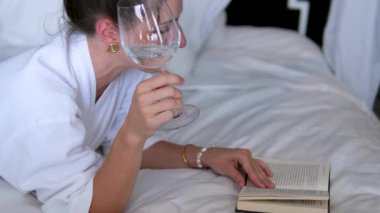 Kadın yatağında e-kitap okuyor, kadın karnının üstünde yatan bir kitap okuyor. Kar beyazı yatak, bornoz, pahalı otel, tatil, bilgi.