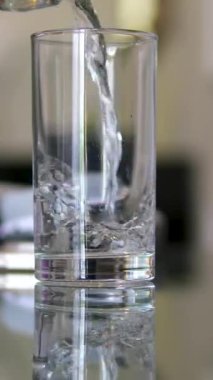 Güneşli ahşap mutfak masasında bardağa dökülen temiz su ve arka planda gri duvar, ağır çekim. Yüksek kalite 4k görüntü