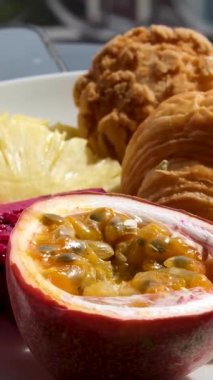 Bir çay kaşığı güneşte, beyaz bir tabakta, tropikal meyve seti, mandalina, tutku meyvesi, ejderha meyvesi, ananas ve kruvasan ile ye. Yüksek kalite 4k görüntü