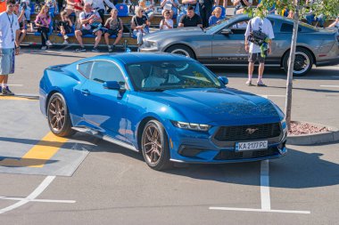 Açılı mavi bir Ford Mustang GT, gün ışığı altında bir yola park etmiş, etrafı yayalar, diğer arabalar, ağaçlar ve şehir merkezindeki potansiyel banklarla çevrili. Sahneleme veya Kyiv, Ukrayna - 21 Eylül 2025