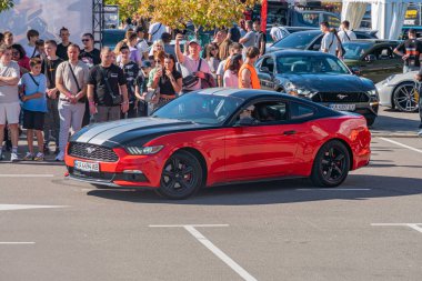 Bir araba fuarında kırmızı Ford Mustang, kalabalık hayranlığı, açık hava ayarı, parlak bitiş, gündüz aydınlatma, yarı resmi kıyafetler, diğer arabalar, sırt çantaları, el çantaları, Kyiv, Ukrayna - 21 Eylül 2025