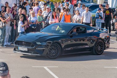 Açık kaputlu siyah bir Ford Mustang spor arabasının halka açık bir toplantıda görüntüsü, performans modifikasyonları gösteriliyor. Aracı çevreleyen ve I Kyiv, Ukrayna - 21 Eylül 2025