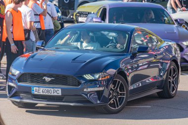 Mavi Ford Mustang GT güneşli bir sokakta, ön planda, parlak tasarım, aerodinamik şekil, parıldayan kaporta, arka planda seyirci ve araç kalabalığı, Kyiv, Ukrayna - 21 Eylül 2025
