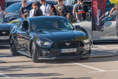 Bir araba fuarında siyah Ford Mustang, parlak yüzeyi ile çeşitli giysi ortamlarında insanlar, muhtemelen gündüz vakti park yeri I Kyiv, Ukrayna - 21 Eylül 2025