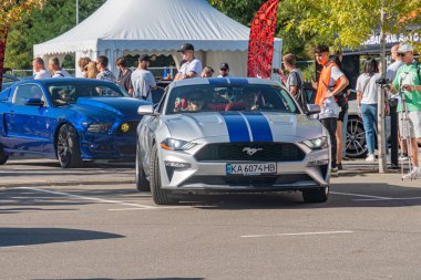 Açık hava araba fuarı veya sergisi, parlak güneş ışığı, iki farklı Ford Mustang mavi, siyah çatı beyaz, mavi yarış çizgileri, parlak bitiş, parlak tasarım, canlı kolo Kyiv, Ukrayna - 21 Eylül 2025