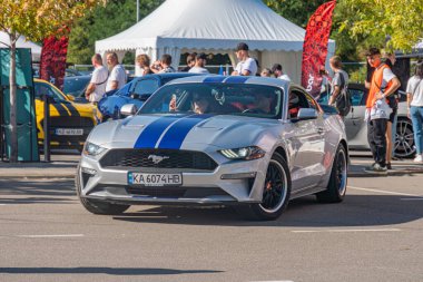 Açık hava arabalarında mavi çizgili gümüş Ford Mustang, arka planda Kyiv, Ukrayna 'da diğer arabaların arasında 21 Eylül 2025' te özel siyah jantlar, doğal ışık altında canlı renkler sergiliyor.