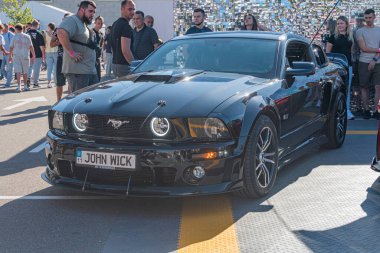 Bir araba fuarında sergilenen siyah Ford Mustang, muhtemelen yarış pistlerini taklit eden sarı ve beyaz çizgilerle özelleştirilmiştir. Araba, büyük çok dilli Kyiv, Ukrayna - 21 Eylül 2025