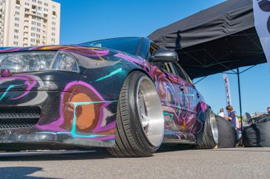 Alacakaranlıkta grafiti boyalı bir arabanın olduğu sokak sahnesi, çok balkonlu bir binanın önüne park edilmiş. Geniş tekerlekler, Kyiv, Ukrayna 'da 21 Eylül 2025' te kurulmuş bir gösteri veya stil modifikasyonları çadırına işaret ediyor.