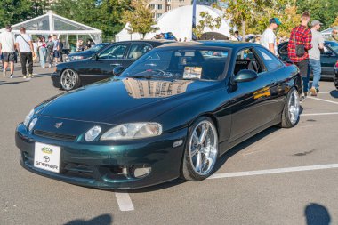 Siyah bir Toyota GT86 spor arabası, beyaz bir çadırın yanında, insanların günlerini geçirdikleri bir şehir ya da yarı şehir merkezinde park halindedir.