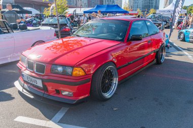 Özel yapım gövdesi olan kırmızı bir BMW E30 coupe, yan etekler, arka spoiler ve alçak binici duruşu nedeniyle asfalt yüzeyine park etmiş. 21 Eylül 2025