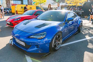 Merkez Mavi Nissan GT-R oto fuarında, canlı kırmızı spor araba solda, açılı modern araç sağda, arka planda bulanık figürler, bulutlu gün, canlı renkler Kyiv, Ukrayna - 21 Eylül 2025