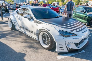 Siyah ve gri grafikli beyaz spor araba, arka planda ikinci araçlar gösteren bir arabanın asfalt yüzeyine park edilmiş, insanlar doğal ışıklandırma, Kyiv, Ukrayna - 21 Eylül 2025