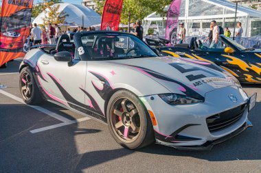 Pembe, mavi ve sarı alev desenli gri Mazda Miata 'nın yer aldığı canlı bir açık hava sahnesi. Araba bir çadır yapının yanına park edilmiş ve ek olarak Kyiv, Ukrayna - 21 Eylül 2025