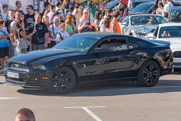 Siyah Ford Mustang GT, halka açık bir etkinlikte yatay olarak park etmiş durumda, muhtemelen bir araba şovu. Farklı insanlardan oluşan kalabalık, doğal ışıklandırma ve Kyiv, Ukrayna - 21 Eylül 2025