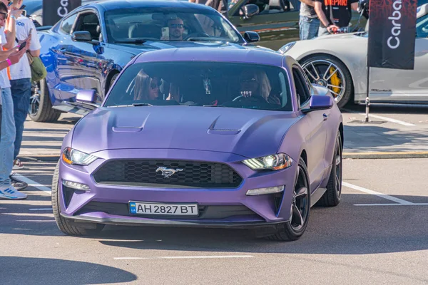Canlı mor bir Ford Mustang, aerodinamik özellikler sergiliyor, açık hava otomobil etkinliklerinde işaretli beton bir yüzeye park edilmiş durumda. Seyirciler toplanıyor, dikkatleri d Kyiv, Ukrayna - 21 Eylül 2025