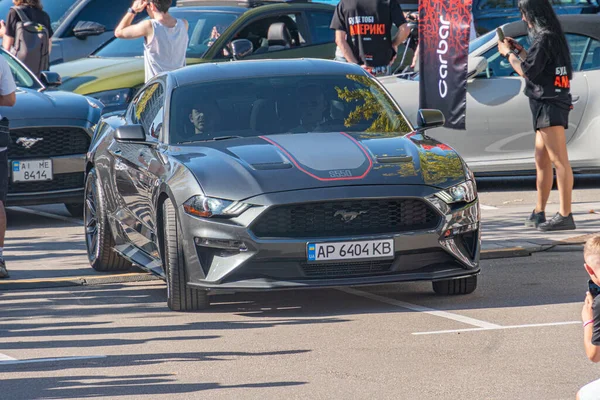 Koyu renkli bir Ford Mustang spor arabası bir araba fuarında sergileniyor. Arabanın çizgili bir çizgisi, özel çıkartması ve aerodinamik bir tasarımı var. Üç fotoğraf Kyiv, Ukrayna 'yı gösteriyor.
