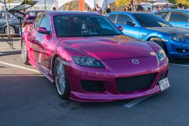 Pembe Mazda Miata yol kenarına park etmiş, tasarım ve spoiler sunuyor. Akşamın geç saatlerinde aydınlatma, uzun gölgeler ve yumuşak ışıltı yayıyor. Ukrayna, Kyiv 'deki diğer araçlar - 21 Eylül 2025