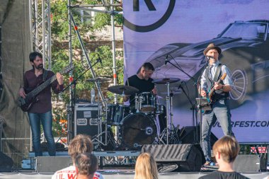 Parlak sahne ışıkları altında bir şarkıcı, davulcu ve bas gitaristin sahne aldığı açık havada canlı müzik performansı. Seyirciler Ukrayna 'nın başkenti Kyiv' de 21 Eylül 2025 'de sergilenen etkinlik logosundan izliyorlar.